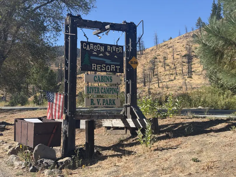 The resort's roadside sign on the highway: Carson River Resort, over Cabins, River Camping and R.V. Park, with a flag and a dry, burned hillside behind.