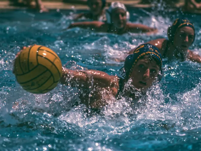 A water polo player lunges for a yellow ball in a burst of spray, teammates in capped helmets behind, in the faded color of a late-1970s photograph.